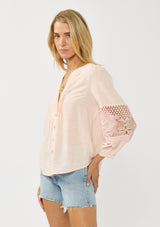 [Color: Blush] A side facing image of a blonde model wearing a pink blouse with a button front, split v-neckline, pintuck details, and a lace applique on the sleeves. A warm weather style part of the resort collection. 