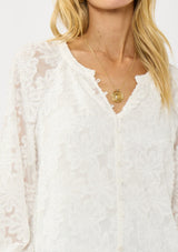 [Color: White] A detail image of a blonde model wearing a white blouse with a clipped floral design, fuzzy texture, button front, and long sheer sleeves. An everyday warm weather style. 