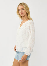 [Color: White] A side facing image of a blonde model wearing a white blouse with a clipped floral design, fuzzy texture, button front, and long sheer sleeves. An everyday warm weather style. 