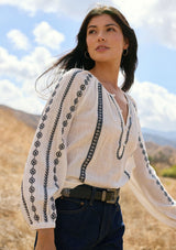 [Color: White/Black] A side facing image of a brunette model wearing a white cotton top with black embroidered details. A classic boho top with a split v-neckline, button front, and long sleeves.  
