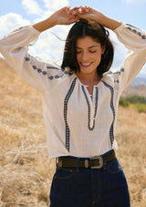[Color: White/Black] A front facing image of a brunette model wearing a white cotton top with black embroidered details. A classic boho top with a split v-neckline, button front, and long sleeves.  