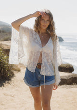 [Color: Natural] A front facing image of a blonde model at the beach wearing a beige sheer lace cover up with floral embroidery. Designed with wide kimono sleeves and a delicate tie front detail for easy styling. Perfect for the beach or vacation styling. 