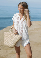[Color: White/Dusty Rose] A front facing image of a blonde model at the beach wearing a white short sleeve top with pink embroidery, lace inserts, and a split v-neckline. Styled with matching bottoms. 