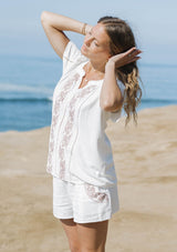 [Color: White/Dusty Rose] A front facing image of a  blonde model at the beach wearing a white short sleeve top with pink embroidery, lace inserts, and a split v-neckline. Styled with matching bottoms. 