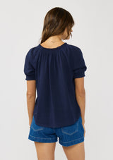 [Color: Eclipse Blue] A back facing image of a brunette model wearing a lightweight dark blue top with puff sleeves and a split neckline with tassel ties. 