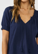 [Color: Eclipse Blue] A detail image of a brunette model wearing a lightweight dark blue top with puff sleeves and a split neckline with tassel ties. 