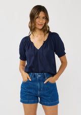 [Color: Eclipse Blue] A front facing image of a brunette model wearing a lightweight dark blue top with puff sleeves and a split neckline with tassel ties. 