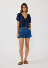 [Color: Eclipse Blue] A front facing image of a brunette model wearing a lightweight dark blue top with puff sleeves and a split neckline with tassel ties. 