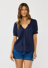 [Color: Eclipse Blue] A front facing image of a brunette model wearing a lightweight dark blue top with puff sleeves and a split neckline with tassel ties. 