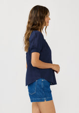 [Color: Eclipse Blue] A side facing image of a brunette model wearing a lightweight dark blue top with puff sleeves and a split neckline with tassel ties. 