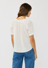 [Color: White] A back facing image of a brunette model wearing a lightweight white top with puff sleeves and a split neckline with tassel ties. 