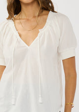 [Color: White] A detail image of a brunette model wearing a lightweight white top with puff sleeves and a split neckline with tassel ties. 