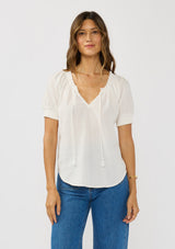 [Color: White] A front facing image of a brunette model wearing a lightweight white top with puff sleeves and a split neckline with tassel ties. 