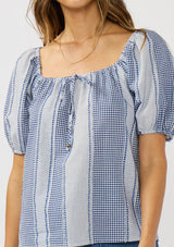 [Color: Navy/White] A detail image of a brunette model wearing a blue and white checkered puff sleeve top with an elastic round neckline. 
