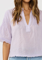 [Color: Lilac] A detail image of a brunette model wearing a light purple top with short sleeves and pintuck details. A lightweight spring top with a soft wrinkled texture. 