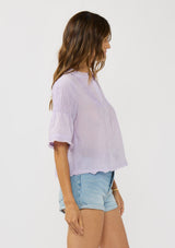 [Color: Lilac] A side facing image of a brunette model wearing a light purple top with short sleeves and pintuck details. A lightweight spring top with a soft wrinkled texture. 