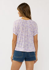 [Color: Natural/Lilac] A back facing image of a brunette model wearing a purple floral top with a short puff sleeves and a round neckline. 