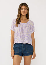[Color: Natural/Lilac] A front facing image of a brunette model wearing a purple floral top with a short puff sleeves and a round neckline. 