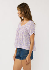 [Color: Natural/Lilac] A side facing image of a brunette model wearing a purple floral top with a short puff sleeves and a round neckline. 