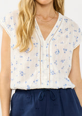 [Color: White/Blue] A detail image of a blonde model wearing a white and blue floral top with a v neckline, short dolman sleeves, and an elastic hem at the back. 