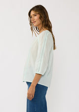 [Color: Mint] A side facing image of a brunette model wearing a sheer mint blouse with lace trims and a hidden button front.