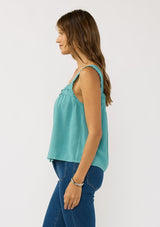 [Color: Teal] A side facing image of a brunette model wearing a bohemian teal tank top with a button front, square neckline, and wide straps with ruffle trims. 