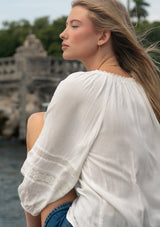 [Color: Off White] A back facing image of a blonde model outside wearing a white boho blouse with embroidered and lace details. 