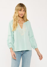 [Color: Dusty Mint] A front facing image of a blonde model wearing a mint green boho blouse with embroidered and lace details. 