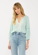 [Color: Dusty Mint] A front facing image of a blonde model wearing a mint green boho blouse with embroidered and lace details. 