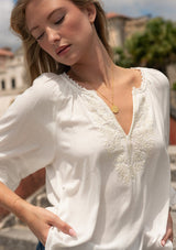 [Color: Off White] A front facing image of a blonde model outside wearing a white boho blouse with embroidered and lace details. 