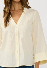 [Color: Eggshell] A detail image of a brunette model wearing a pale yellow blouse with a v-neckline, button front, and wide long sleeves. 