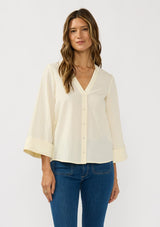 [Color: Eggshell] A front facing image of a brunette model wearing a pale yellow blouse with a v-neckline, button front, and wide long sleeves. 