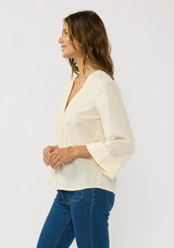 [Color: Eggshell] A side facing image of a brunette model wearing a pale yellow blouse with a v-neckline, button front, and wide long sleeves. 