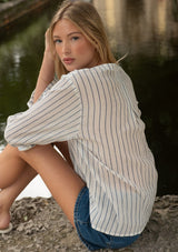 [Color: White/Denim Blue] A sitting image of a blonde model wearing a blue and white striped blouse with a v neckline and long sleeves. 