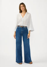 [Color: White/Denim Blue] A side facing  image of a brunette model wearing a blue and white striped blouse with a v neckline, button front, and long wide sleeves. Styled tucked in with dark wash denim 