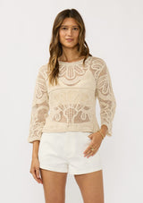 [Color: Natural] A front facing image of a brunette model wearing a beige crochet knit top with long sleeves and a round neckline. Styled with white shorts for a beach ready look. 