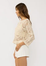 [Color: Natural] A side facing image of a brunette model wearing a beige crochet knit top with long sleeves and a round neckline. Styled with white shorts for a beach ready look. 