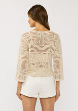 [Color: Natural] A back facing image of a brunette model wearing a beige crochet knit top with long sleeves and a round neckline. Styled with white shorts for a beach ready look. 