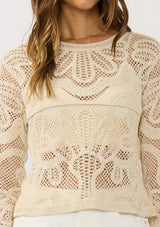 [Color: Natural] A detail image of a brunette model wearing a beige crochet knit top with long sleeves and a round neckline. Styled with white shorts for a beach ready look. 