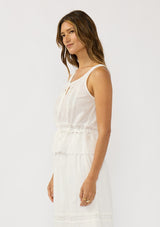 [Color: White] A side facing image of a brunette model wearing a white cotton tank top with an eyelet trimmed hem, a drawstring waist, and a round neck with tie detail. Styled with a matching white maxi skirt. 