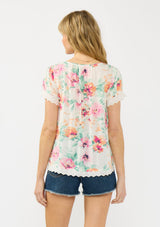 [Color: Ivory/Fuchsia] A back facing image of a blonde model wearing a spring floral top with an eyelet trim, a scoop neckline, and a button front. 
