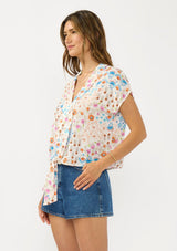 [Color: Ivory/Orange] A side facing image of a brunette model wearing an ivory and orange floral boho top with short sleeves, a v neckline, and a tie front detail.