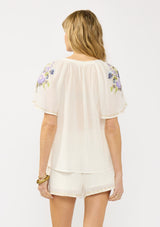 [Color: Natural/Lilac] A back facing image of a blonde model wearing an bohemian embroidered short sleeve top with ties and a button front. 