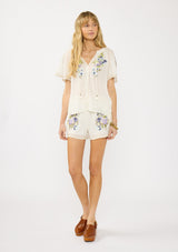 [Color: Natural/Lilac] A front facing image of a blonde model wearing an bohemian embroidered short sleeve top with ties and a button front. 