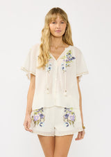 [Color: Natural/Lilac] A front facing image of a blonde model wearing an bohemian embroidered short sleeve top with ties and a button front. 
