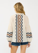 [Color: Natural/Denim] A back facing image of a blonde model wearing a beige and blue jacquard blouse with a zigzag design, long voluminous sleeves, and a split v neckline. 