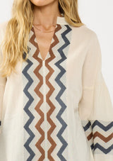 [Color: Natural/Denim] A detail image of a blonde model wearing a beige and blue jacquard blouse with a zigzag design, long voluminous sleeves, and a split v neckline. 