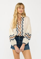 [Color: Natural/Denim] A front facing image of a blonde model wearing a beige and blue jacquard blouse with a zigzag design, long voluminous sleeves, and a split v neckline. 