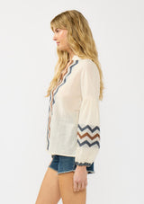 [Color: Natural/Denim] A side facing image of a blonde model wearing a beige and blue jacquard blouse with a zigzag design, long voluminous sleeves, and a split v neckline. 