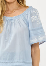 [Color: Light Blue] A detail image of a blonde model wearing a light blue embroidered top with short sleeves and a scoop neckline with ruffle trim.